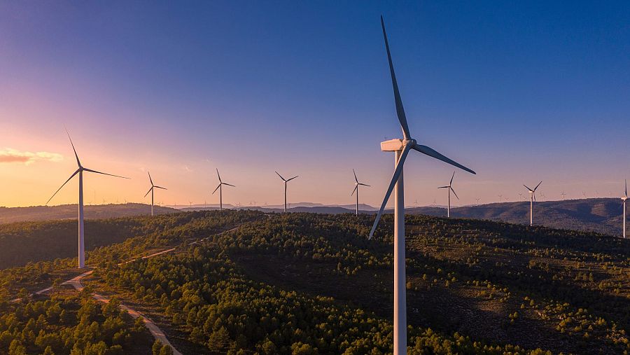 Impresionante paisaje de un parque eólico al amanecer o atardecer. Aerogeneradores blancos sobre colinas con vegetación y un camino.  Cielo con tonos rosados y azules.