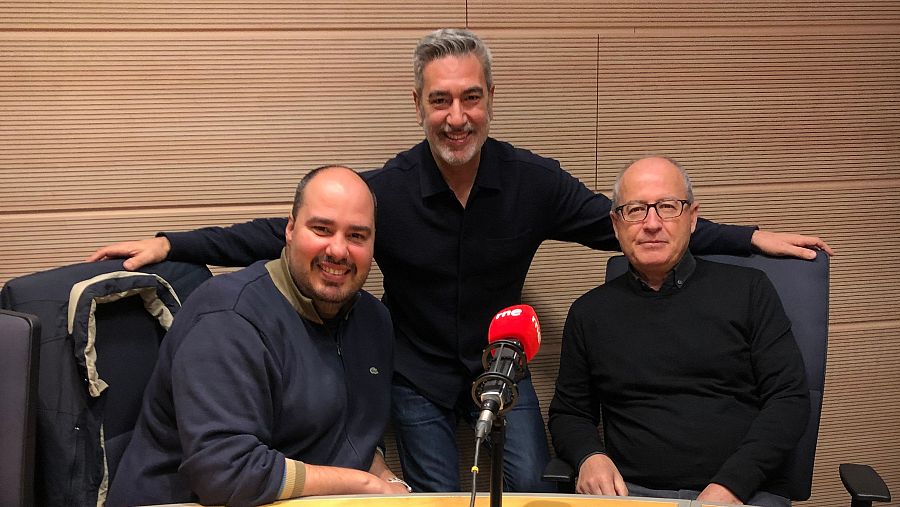 Tres hombres sentados alrededor de una mesa en un estudio de radio con un micrófono; uno sonríe, otro sonríe y el tercero tiene una expresión neutra.