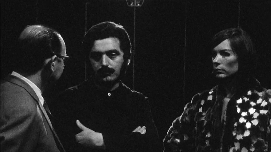 Imagen en blanco y negro: Paco Rabanne, con bigote y camisa oscura, conversa con un hombre en traje y una mujer con abrigo claro. Fondo oscuro con líneas verticales.