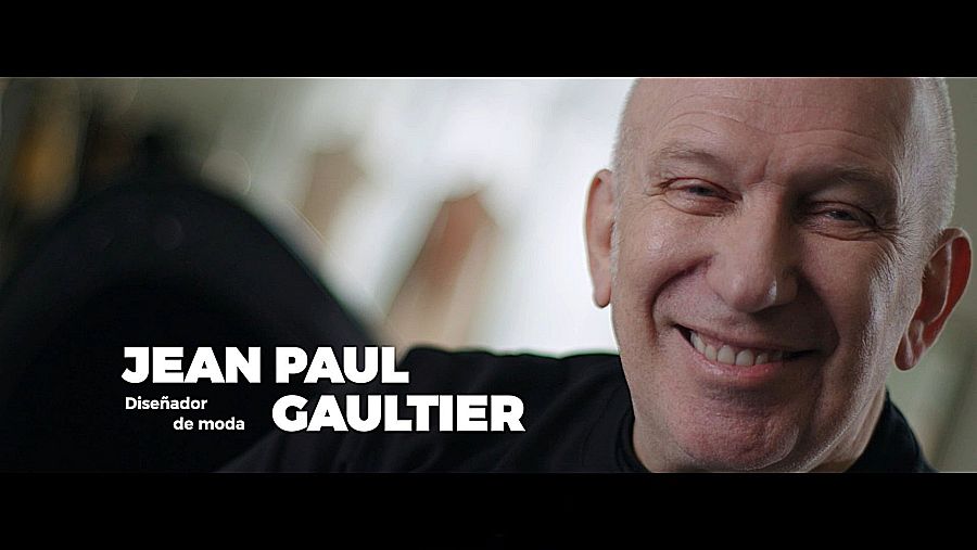 Jean Paul Gaultier, diseñador de moda, sonríe en un primer plano de fotograma de documental. Texto superpuesto confirma su identidad.