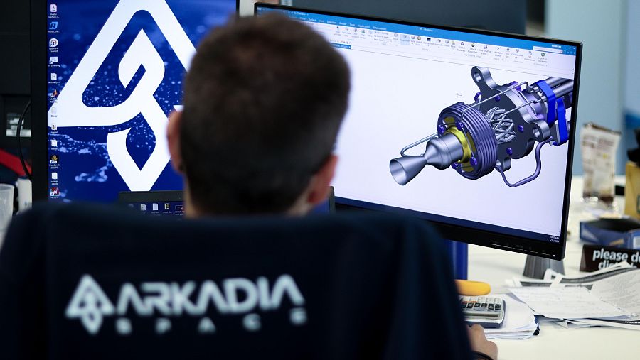 Hombre trabajando en ordenador con diseño 3D de pieza mecánica en pantalla.  Escritorio con papeles y calculadora. Logo Arkadia visible.