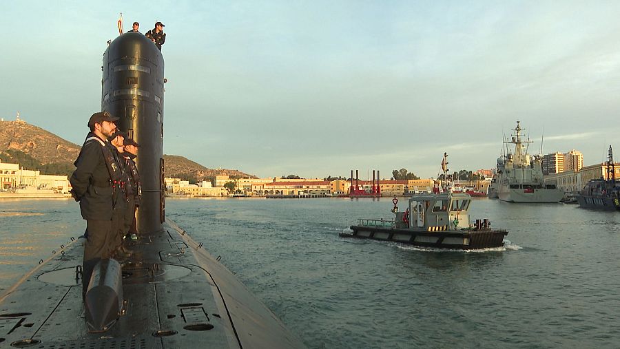 Tripulantes en la cubierta y torre de mando de un submarino, con un remolcador cercano y la línea de costa al fondo, iluminada por la luz del atardecer.
