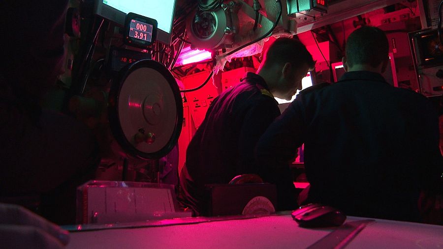 Cámara de mando iluminada en rojo, dos tripulantes trabajando en consolas y pantallas que muestran datos numéricos.  Se ven equipos electrónicos, un ratón y una regla.