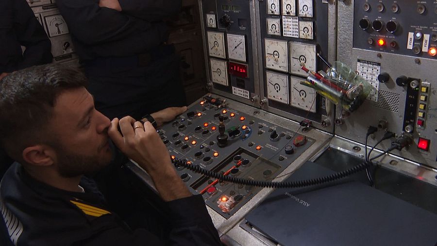 En la sala de máquinas de un submarino, un hombre con uniforme oscuro opera un panel de control con numerosos botones, luces y medidores, usando un dispositivo de comunicación.