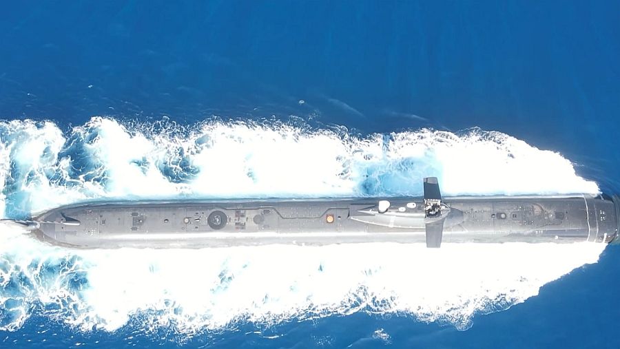 Vista cenital de un submarino gris navegando en aguas oscuras; se aprecian detalles como escotillas y una estela blanca.