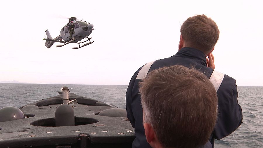 Tripulantes del submarino, con uniformes oscuros, observan un helicóptero en maniobra sobre el mar.  Se ve parte de la estructura del submarino y el helicóptero en vuelo estacionario.