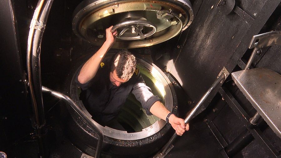 Tripulante en uniforme sale por escotilla circular de submarino; usa rueda y barra metálica para apoyarse; interior de la escotilla verde claro.