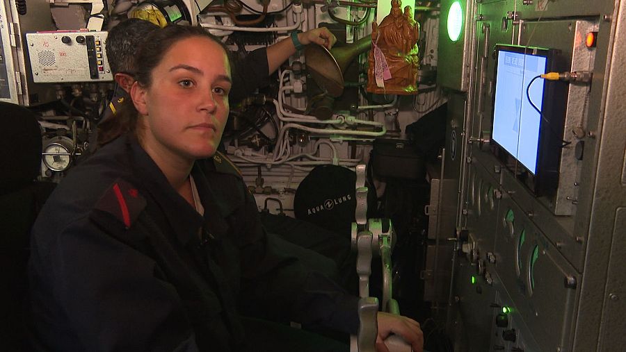 Timonel femenina en la cabina de un submarino, frente a paneles de control, indicadores y palancas. Iluminación tenue, ambiente serio.