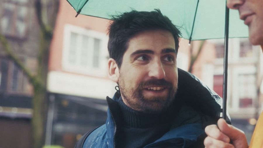 Hombre con barba y chaqueta azul bajo paraguas verde en día lluvioso. Sonríe y mira a la derecha; otra persona aparece parcialmente en la imagen.