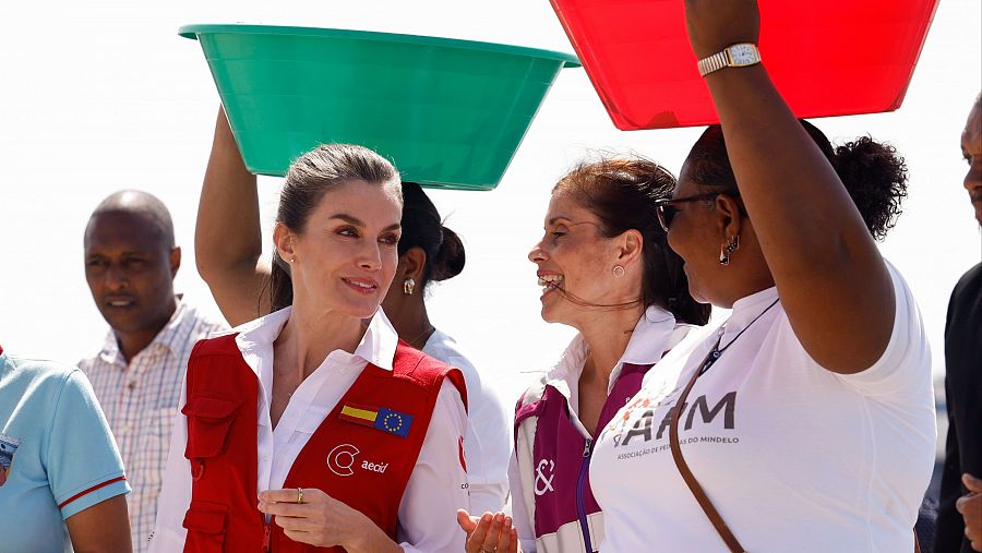 La Reina Letizia, en un chaleco rojo con logos de la Cooperación Española y la UE, interactúa con personas en un mercado, mostrando una actitud cercana y amable.
