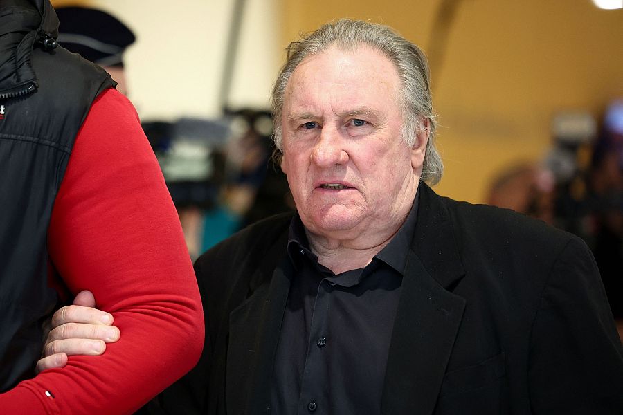 Imagen de Gérard Depardieu llegando a una vista judicial; viste camisa negra y chaqueta oscura, expresión seria.  Al fondo, un tono amarillo claro.