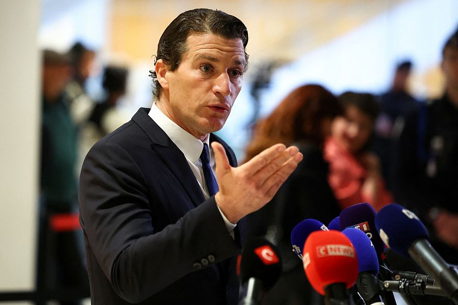 Abogado en traje oscuro y corbata azul, gesticulando mientras habla ante micrófonos en una rueda de prensa.