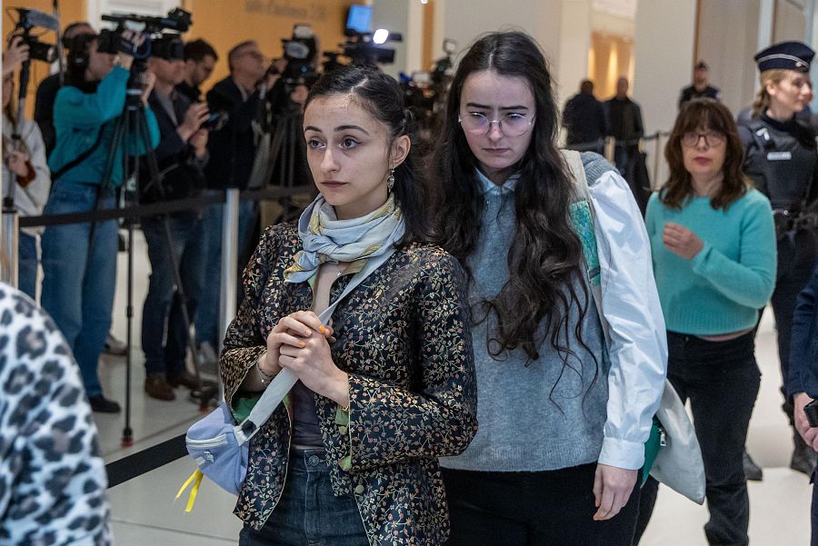 Charlotte Arnould, en primer plano, con ropa oscura y un bolso lila, acompañada de otras mujeres. Fondo con prensa y posible agente de policía.  Escena pública.