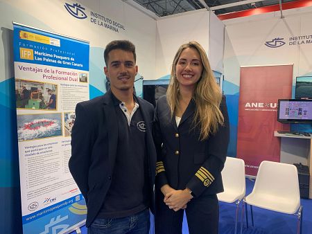 Dos personas en un stand con carteles informativos, uno azul sobre formación profesional y otro rosa con logo ANEXU. Se aprecia un simulador en pantalla.