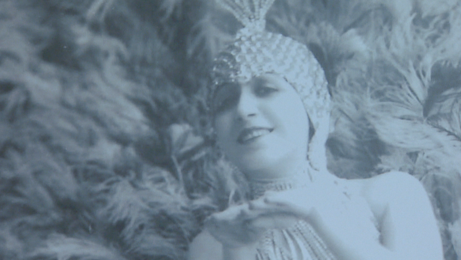 Bailarina de variedades en foto blanco y negro, con tocado de plumas y lentejuelas, y vestido con flecos. Postura de baile o pose.  Imagen histórica.