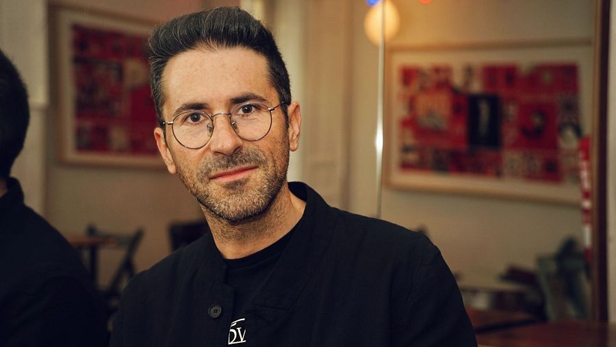 Retrato de Agus Morales en un espacio interior, posiblemente un teatro, con una expresión neutra. Se aprecian cuadros y mobiliario de fondo.