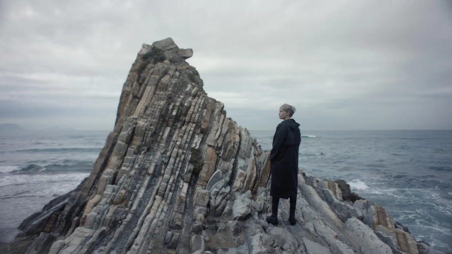 Actriz con abrigo oscuro contemplando un paisaje costero rocoso con mar gris y olas fuertes.