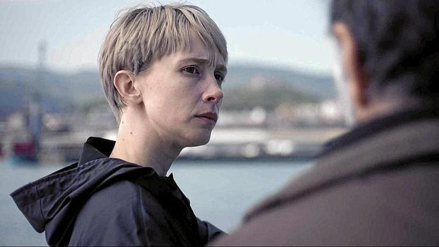 Ingrid García-Jonsson, con chaqueta oscura y pelo rubio platino, en primer plano; al fondo, un puerto desenfocado y parte de otra persona con ropa marrón.  Escena de diálogo.