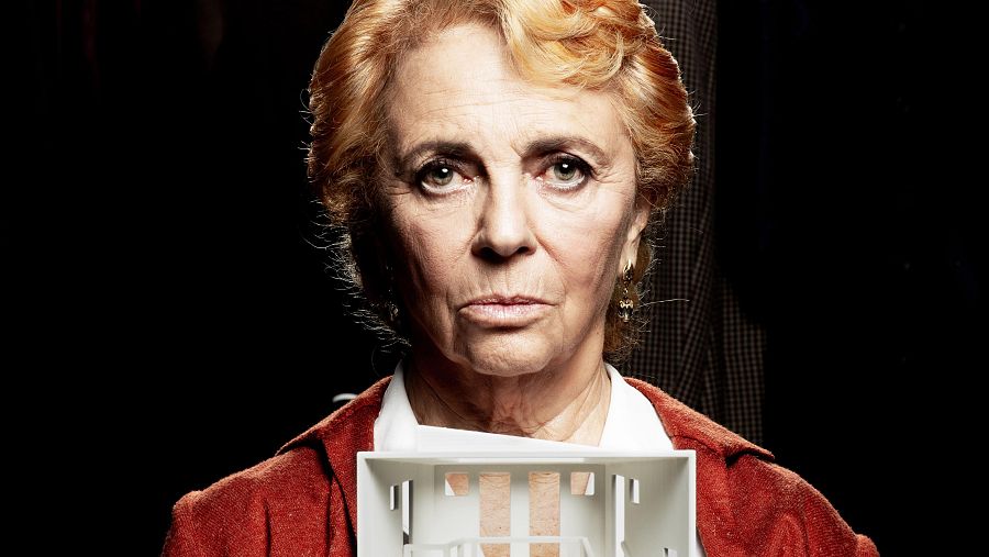 Primer plano de Carmen Machi con expresión seria, chaqueta roja y maqueta blanca de una casa en sus manos. Fondo oscuro.