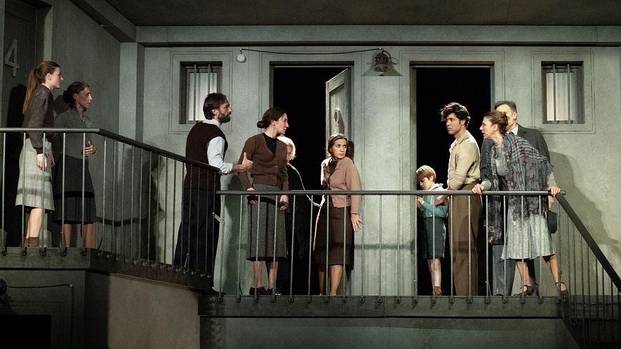 Escena teatral en edificio antiguo con varios personajes (hombres, mujeres y niño) vestidos de época en una escalera. Iluminación tenue, atmósfera dramática. Se ve el número 4 en una puerta.