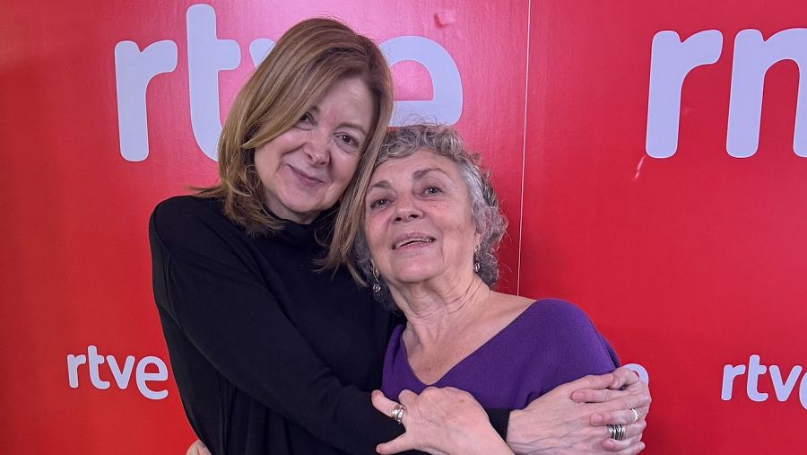 Abrazos entre dos mujeres (rubia con jersey negro y canosa con jersey morado) ante un fondo rojo con logotipos RTVE y RNE.  Posible evento cultural.