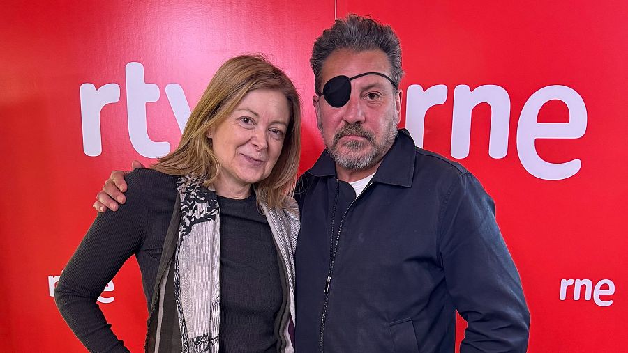 Juan José Millás y una mujer posan ante un fondo rojo con los logos de RTVE y RNE. Él viste chaqueta oscura y parche en el ojo; ella, jersey oscuro y bufanda gris.