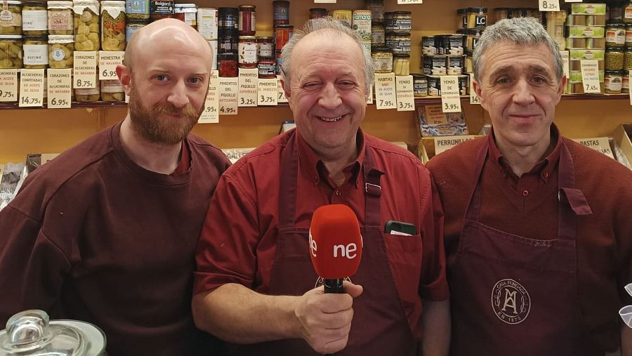 Entrevista en Mantequería Andrés: tres hombres con delantales, uno con micrófono, frente a estanterías con productos envasados y precios visibles.