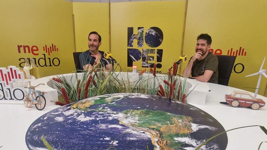 Dos hombres conversan en un podcast de RNE, 'HOPE!', en un estudio con decoración peculiar.  Uno gesticula mientras el otro escucha atentamente.