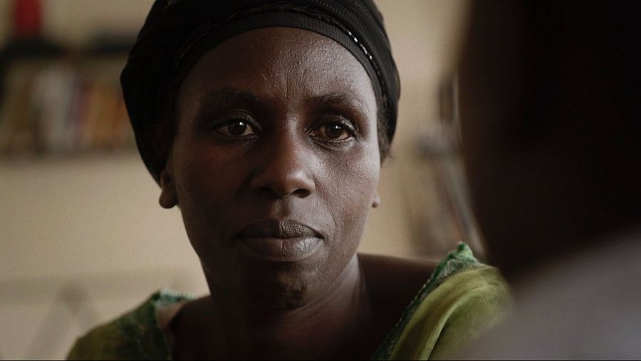 Mujer africana con expresión seria, viste pañuelo negro y ropa verde oliva.  Se observa parte de otra persona desenfocada al fondo.