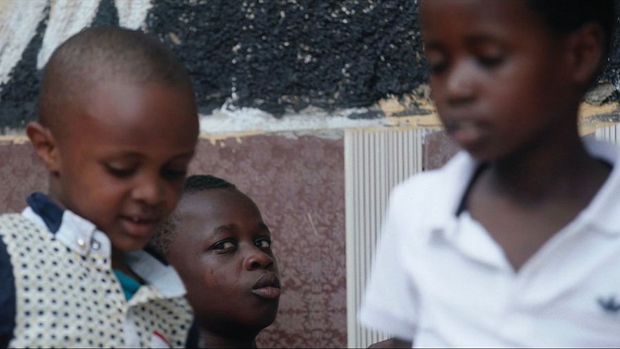 Tres niños, posiblemente ruandeses, posan cerca de una pared con textura.  Uno sonríe, otro mira a la cámara y el tercero mira hacia abajo.