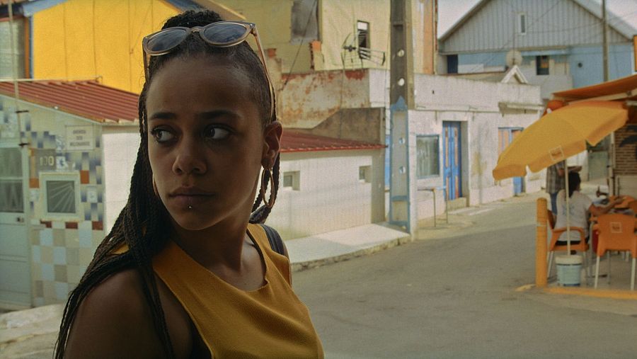 Joven con trenzas y camiseta amarilla mira hacia la derecha en una calle con edificios de colores y una terraza con mesas y sillas naranjas.  Fotograma de 'A nuestros amigos'.