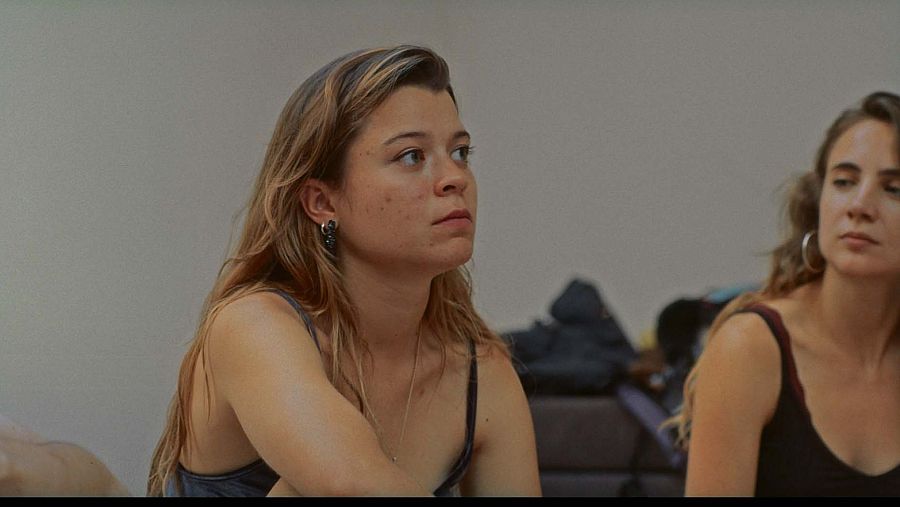 Dos mujeres jóvenes, una rubia y otra castaña, aparecen en un fotograma de película.  Una mira a la derecha, la otra al frente. Fondo borroso con un mueble.