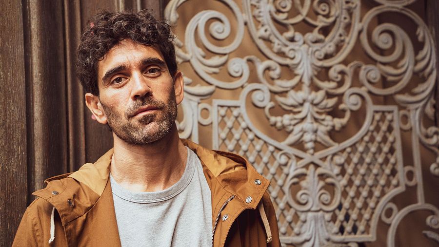 Retrato de Adrián Orr, cineasta, con chaqueta marrón mostaza y camiseta gris, posando ante una puerta ornamentada. Fotografía de Matías Ruedlinger.