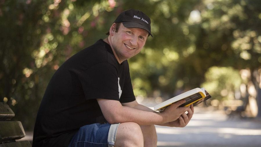 Escritor sentado en un banco de parque, leyendo un libro; viste gorra negra con inscripción, camiseta negra y pantalones cortos vaqueros; expresión relajada.
