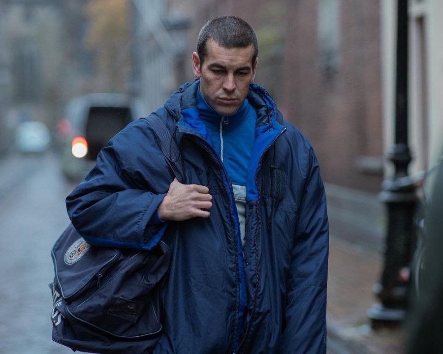 Actor con chaqueta azul marino y bolsa deportiva caminando por una calle mojada. Expresión seria.