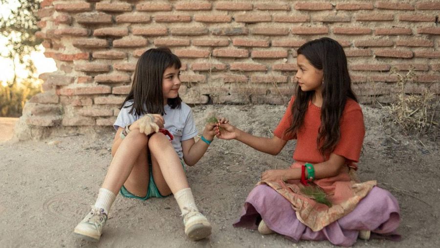 Niñas sentadas en el suelo, cerca de una pared de ladrillo, interactuando; una ofrece una planta a la otra.  Ambiente cálido y tranquilo.
