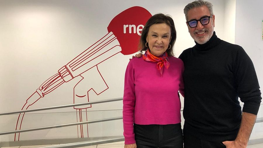 Dos periodistas, una mujer con suéter rosa y un hombre con suéter negro y gafas, posan ante el logo de RNE.