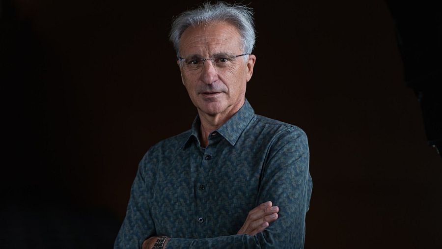 Retrato de un hombre mayor con gafas y camisa estampada, brazos cruzados, sobre fondo negro.