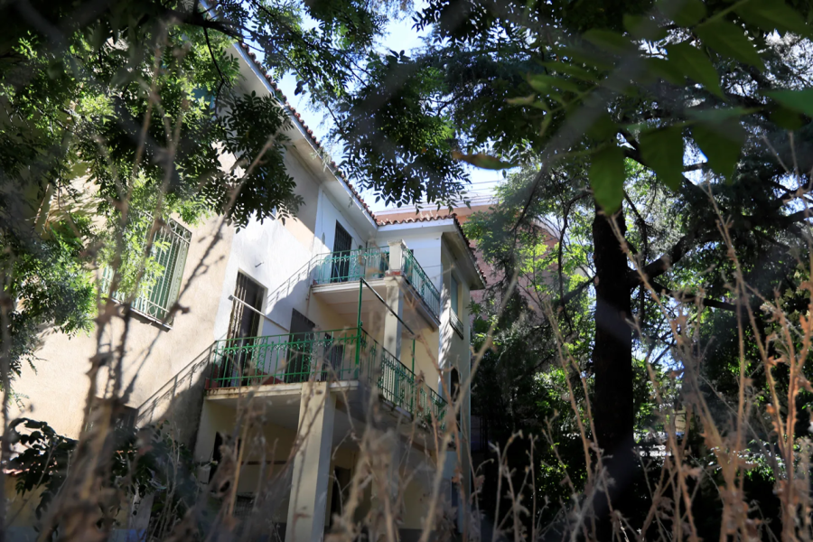 Casa de dos pisos con paredes claras, balcones con barandillas verdes y tejado marrón. Abundante vegetación rodea la construcción.