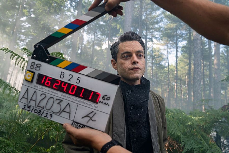 Rami Malek, con abrigo verde oliva, en rodaje forestal. Claqueta: escena B25, toma 88, 13/08/19. Expresión seria.