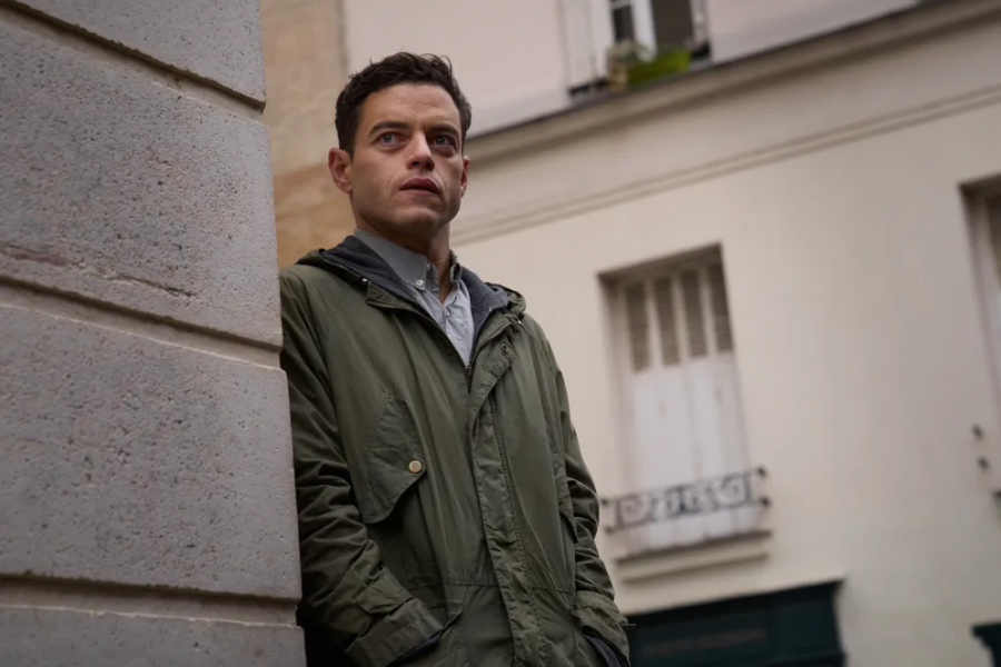 Rami Malek, como Charlie Heller en 'The Amateur', luce una parka verde oliva y una expresión seria mientras se apoya en una pared. Fondo urbano.