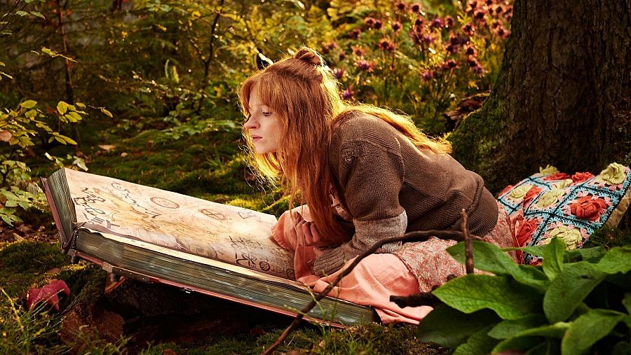 Niña pelirroja leyendo un libro grande con símbolos en un entorno natural soleado. Lleva jersey marrón y falda rosa, sentada sobre una manta.