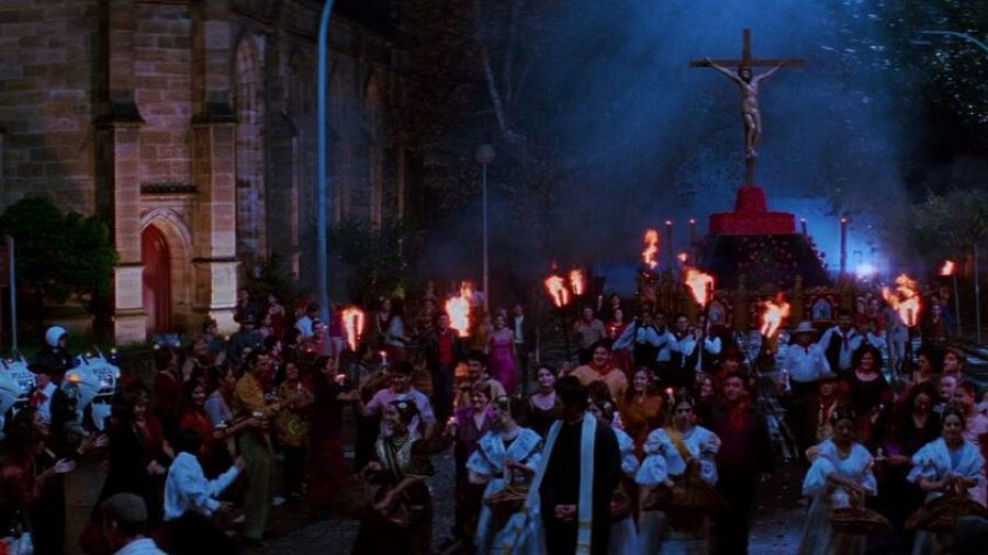 Procesión nocturna con antorchas, figura de Cristo crucificado y multitud festiva. Atmósfera dramática y cálida en una calle con iglesia cercana y presencia policial.