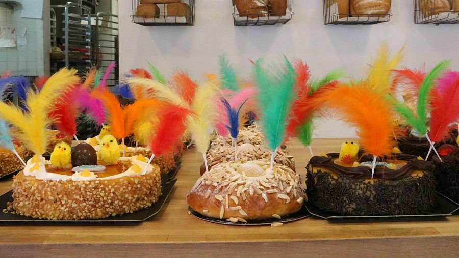 Mones de Pascua con plumas de colores, pollitos amarillos y diversas coberturas (almendras, chocolate, yema).  Exhibidos en bases negras sobre madera.
