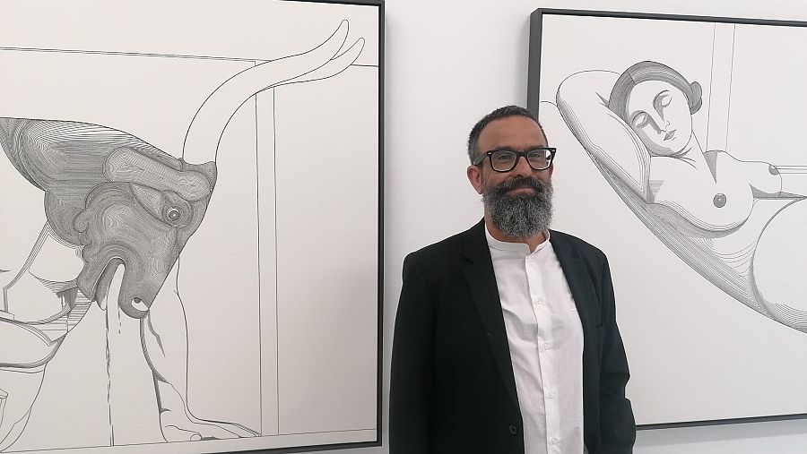 Sergio García, con barba y gafas, entre dos dibujos en blanco y negro enmarcados; uno, un minotauro; el otro, una figura femenina reclinada.