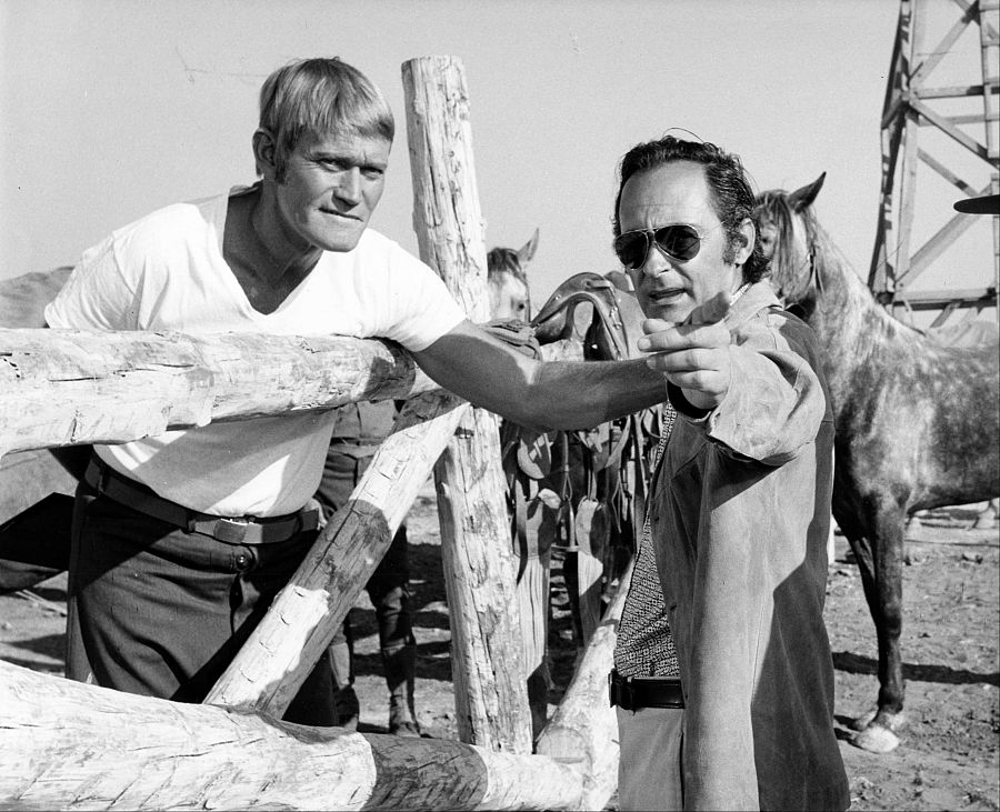 Chuck Connors y Eugenio Martín en una escena de rodaje. Connors viste camiseta y pantalones oscuros; Martín, chaqueta y gafas de sol, parece dirigir.