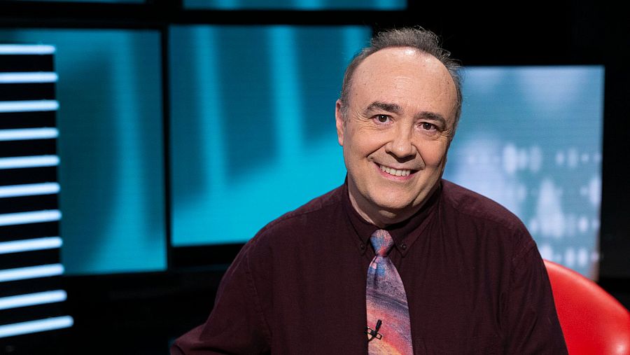 Crítico de cine sonriente en plató de TV con camisa granate y corbata abstracta. Fondo con paneles azules y mobiliario de estudio.