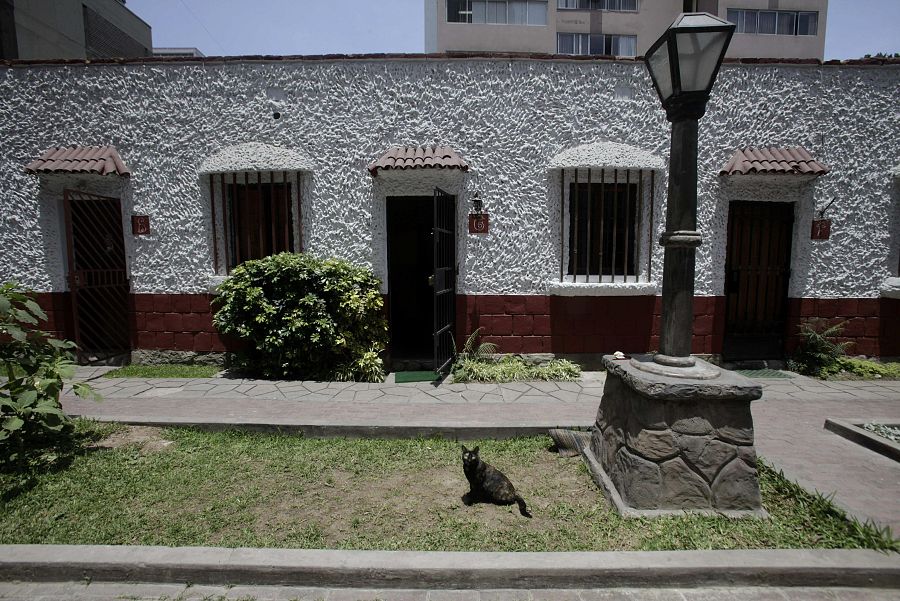 Casa de una planta, paredes blancas y base rojiza, tres puertas con rejas, jardín delantero con farola y un gato negro.