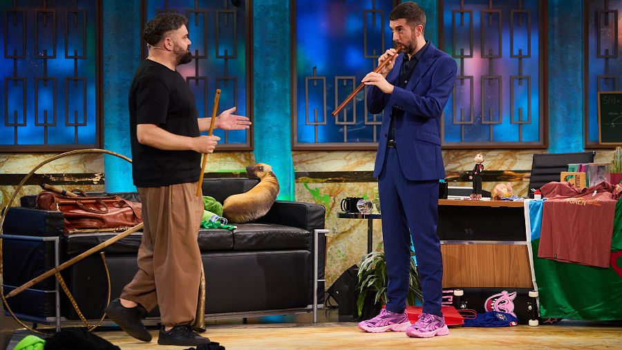 Músico tocando flauta en plató de TV, junto a un presentador y un perro en un sofá. Decoración con ventanas geométricas.