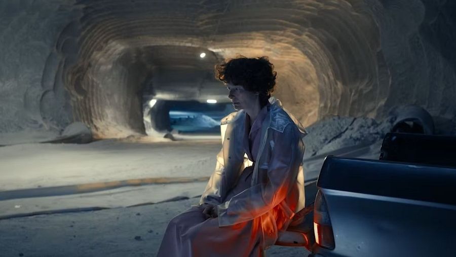 Persona sentada en un túnel rocoso, iluminada por una luz rojiza, con una gabardina clara y un vehículo oscuro cerca.